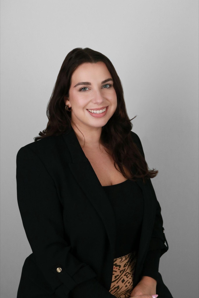 Professional studio headshot of a woman in a black blazer against a neutral gray background